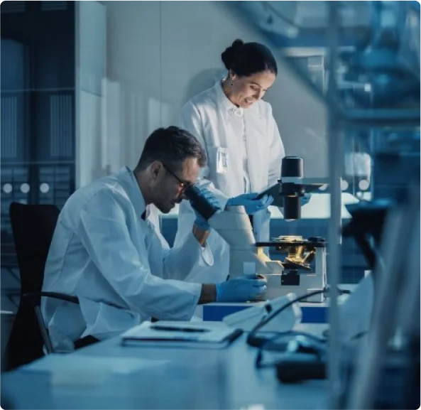 Dois cientistas em um laboratório. Ambos usam jalecos brancos e luvas azuis. Um dos cientistas está olhando através de um microscópio, enquanto a outra está de pé ao lado, observando.